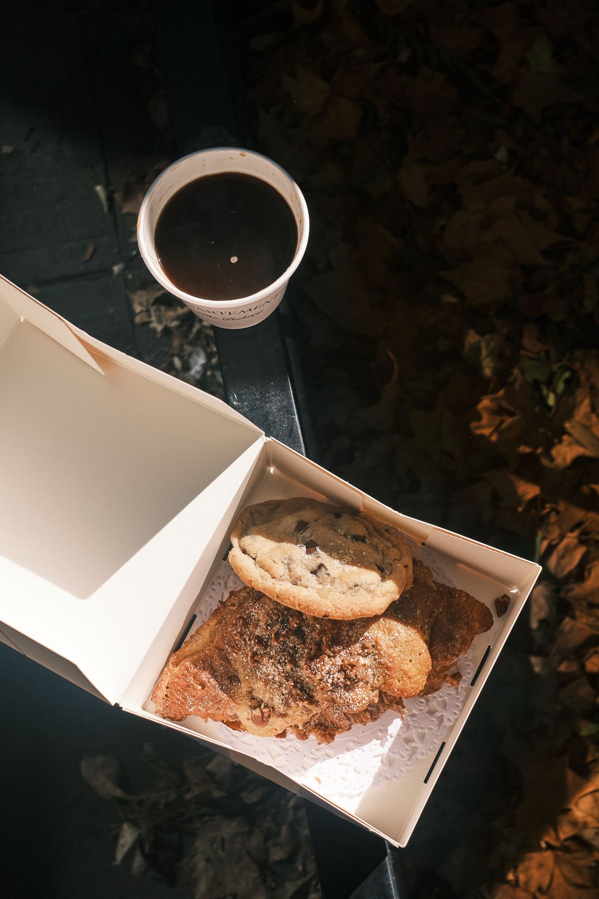 Looking down into an open white bakery box holding a tahini chocolate chip cookie and a flat, sugar-dusted croissant, a cup of black coffee beside it on a dark bench in low autumn sun, with fallen leaves on the ground.