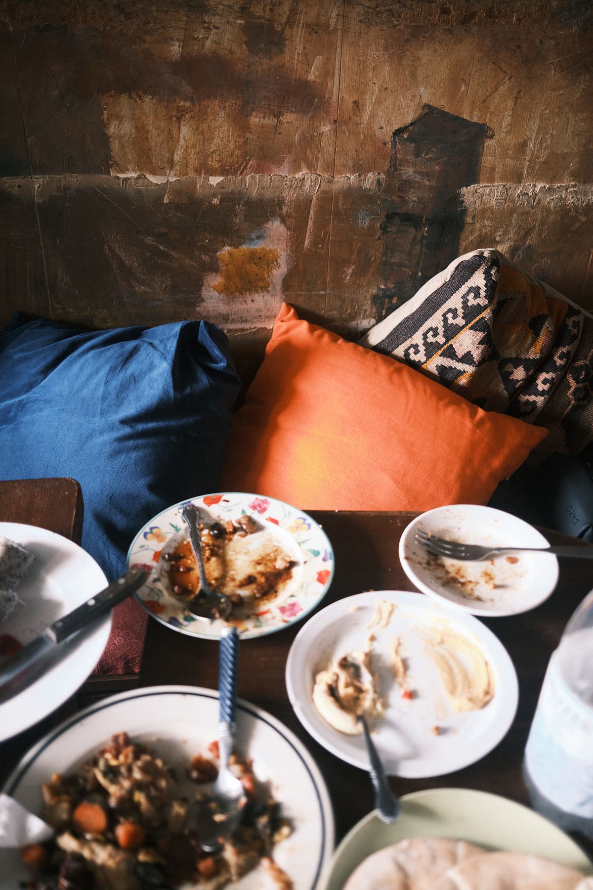 Blue and orange cushions and patterned kilim bolsters propped against a bare, worn plaster wall, with a dark table below holding part-eaten plates of hummus, a stew of carrots and aubergine, flatbread, and a floral-edged plate with a fork resting on it.