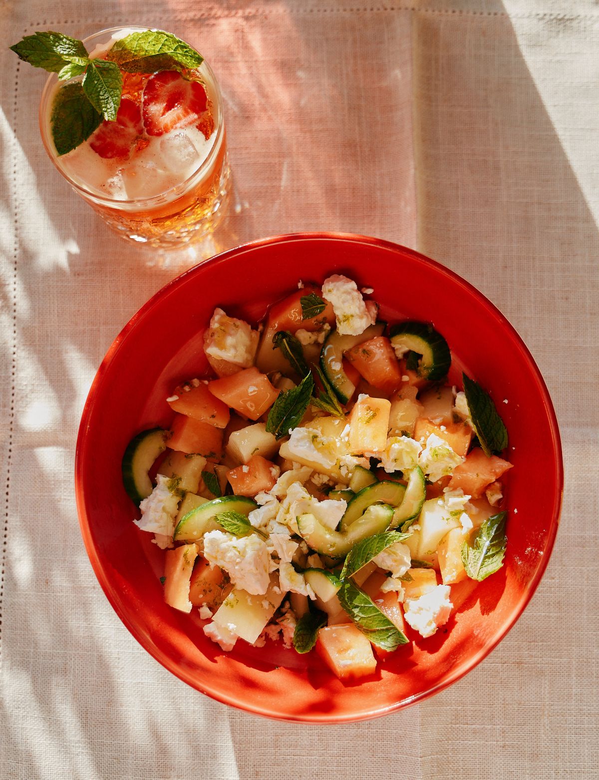 Watermelon, melon, cucumber, feta and mint salad in a red bowl beside an iced drink garnished with strawberry and mint.