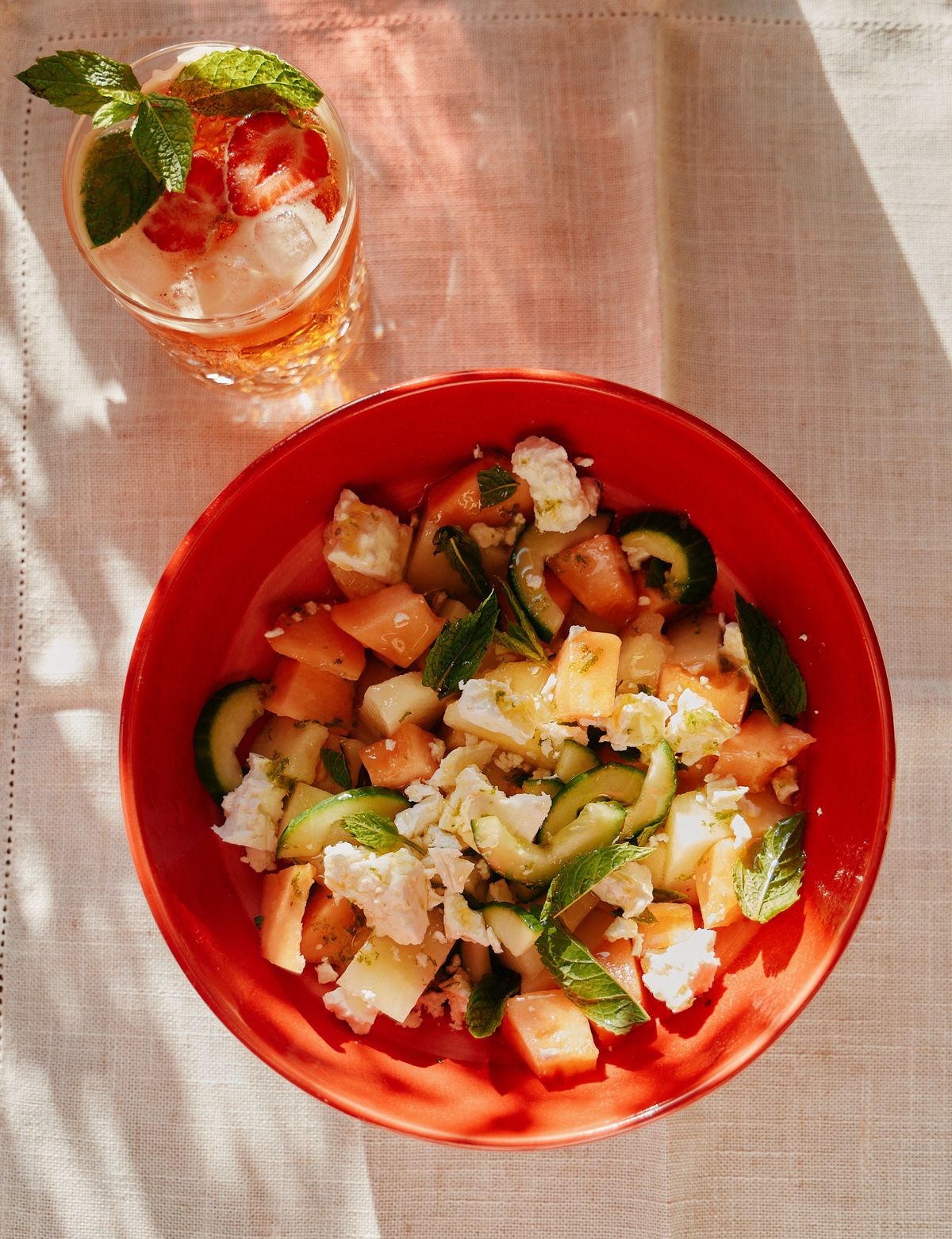 Watermelon, melon, cucumber, feta and mint salad in a red bowl beside an iced drink garnished with strawberry and mint.