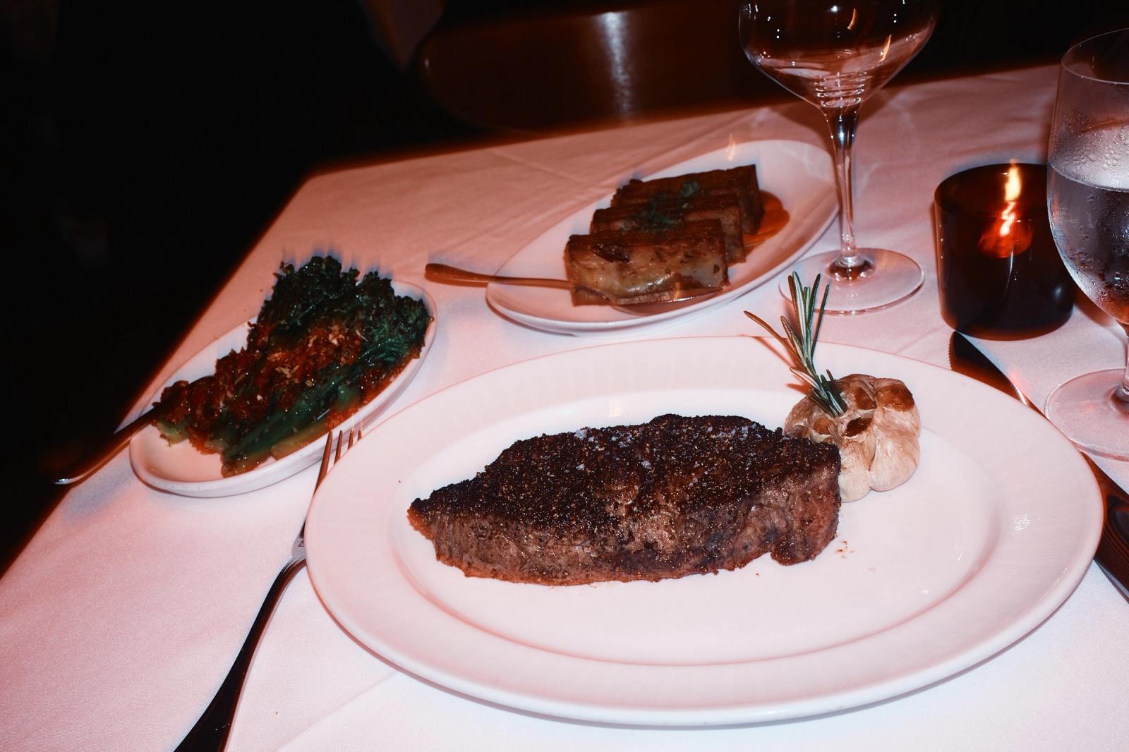 A thick seared strip steak on a white plate with a whole roasted garlic head and a sprig of rosemary, dishes of creamed spinach and bone marrow on the white tablecloth behind, with a wine glass and a lit candle.