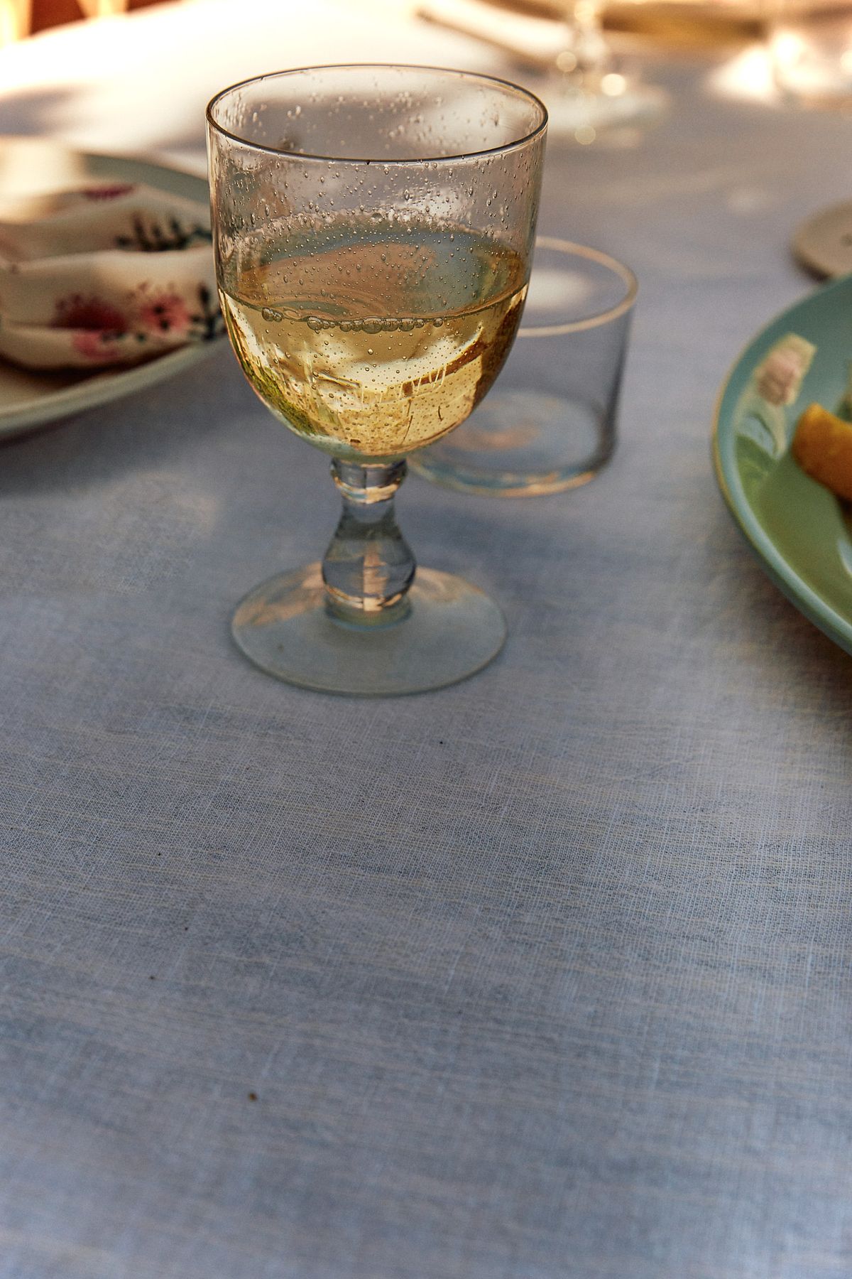 A glass of golden white wine beaded with condensation on a pale blue linen tablecloth, with plates and late afternoon sun around it.