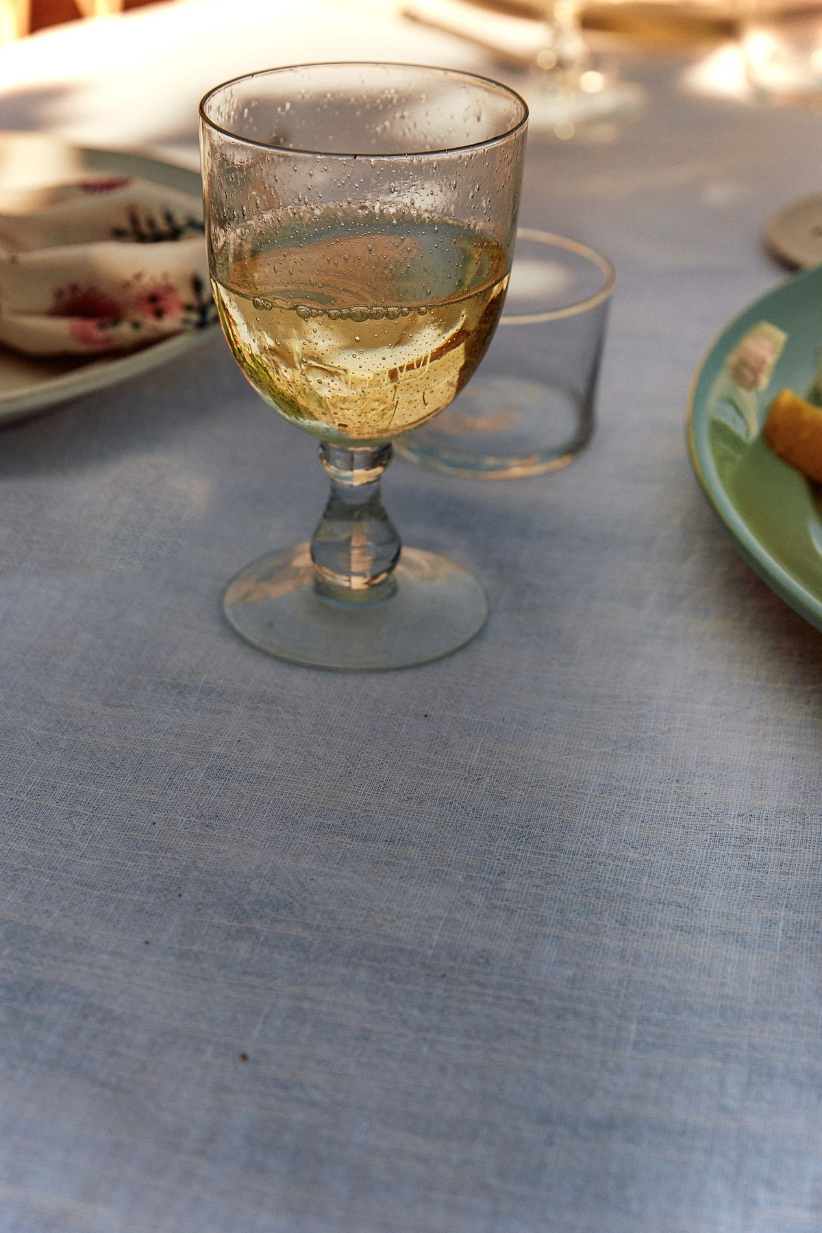 A glass of golden white wine beaded with condensation on a pale blue linen tablecloth, with plates and late afternoon sun around it.