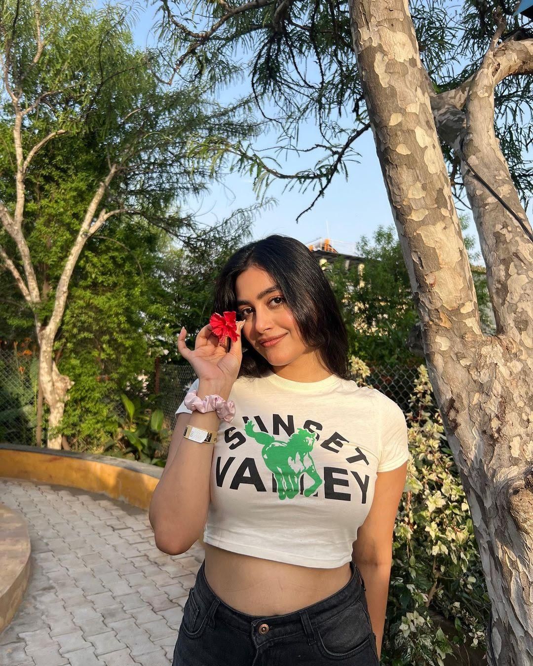 Young woman holding a red hibiscus flower beside a tree on the tree-lined pathway at Rangmanch Farms, an outdoor day outing venue in Gurugram near Delhi