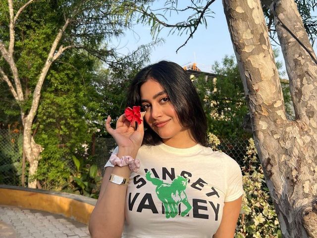 Young woman holding a red hibiscus flower beside a tree on the tree-lined pathway at Rangmanch Farms, an outdoor day outing venue in Gurugram near Delhi