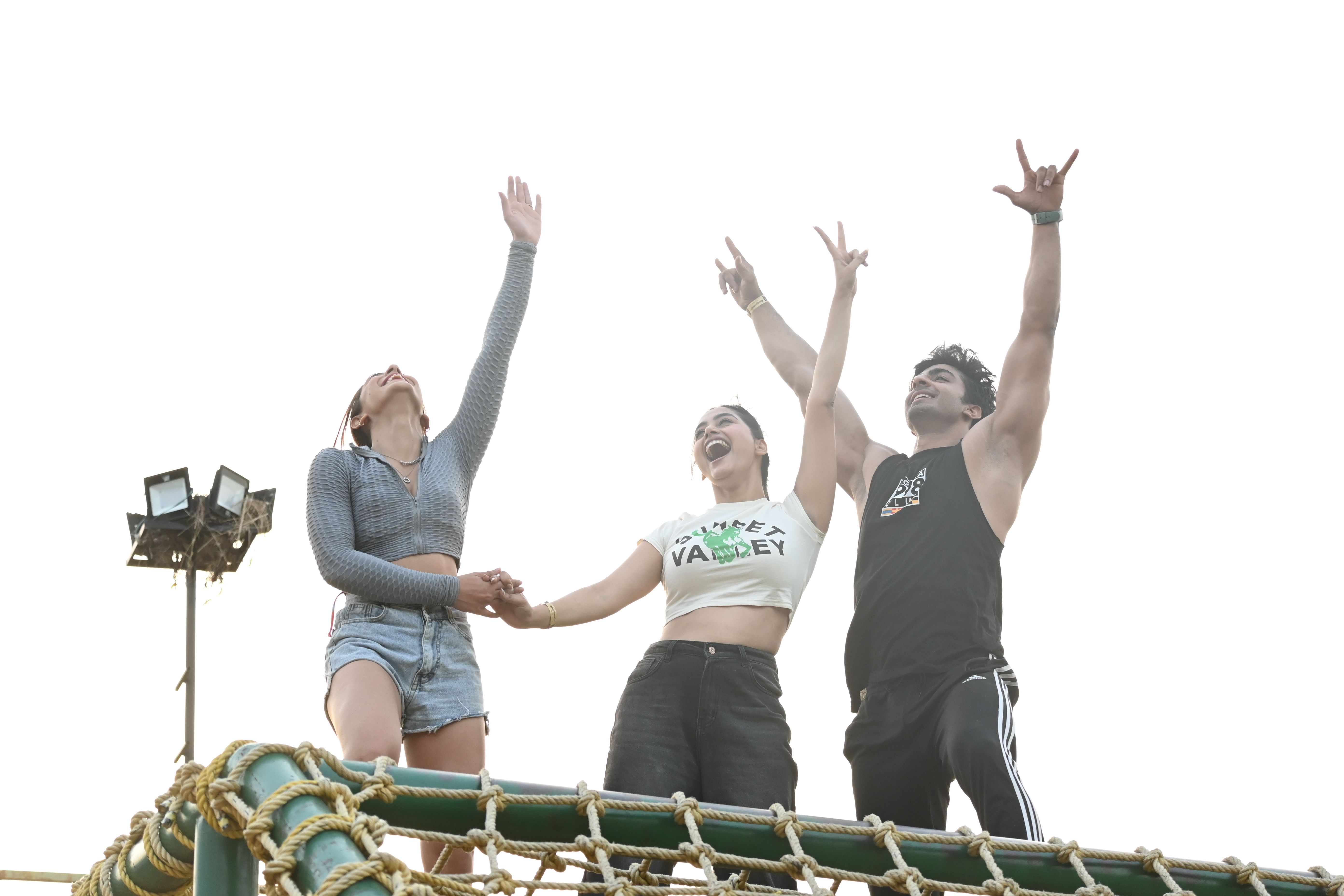 Three friends raising their arms in celebration at the top of a rope climbing obstacle during an outdoor adventure activity at Rangmanch Farms, an outing venue in Gurugram near Delhi