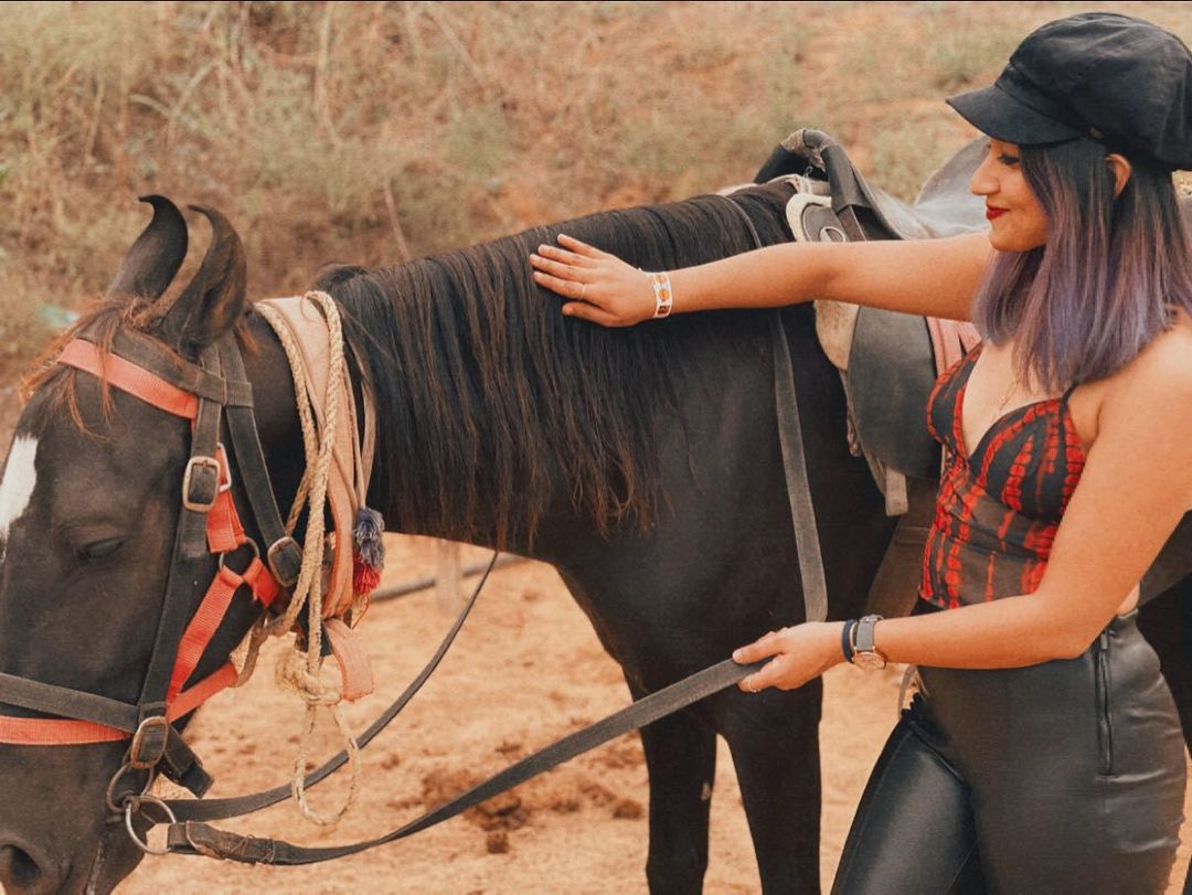 A visitor in a cap petting a saddled black horse on the sandy grounds at Rangmanch Farms, an outdoor day outing venue in Gurugram near Delhi