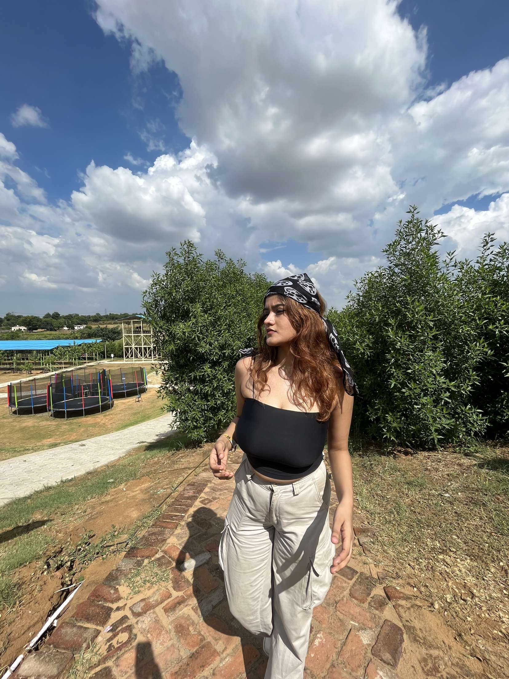 Young woman standing on a brick pathway with trampolines and greenery under a cloudy sky at Rangmanch Farms, an outdoor day outing venue in Gurugram near Delhi