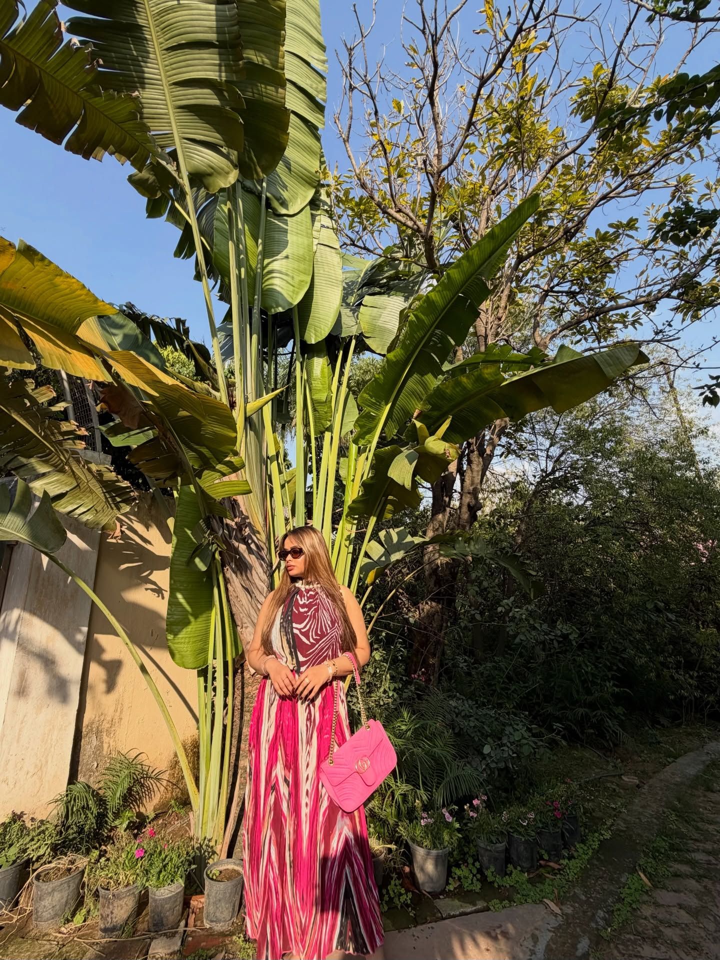Woman in a pink printed dress with a pink handbag standing beside tall banana plants and potted greenery along a garden path at Rangmanch Farms, an outdoor event venue in Gurugram near Delhi