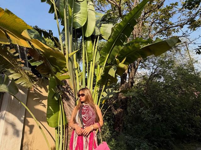 Woman in a pink printed dress with a pink handbag standing beside tall banana plants and potted greenery along a garden path at Rangmanch Farms, an outdoor event venue in Gurugram near Delhi