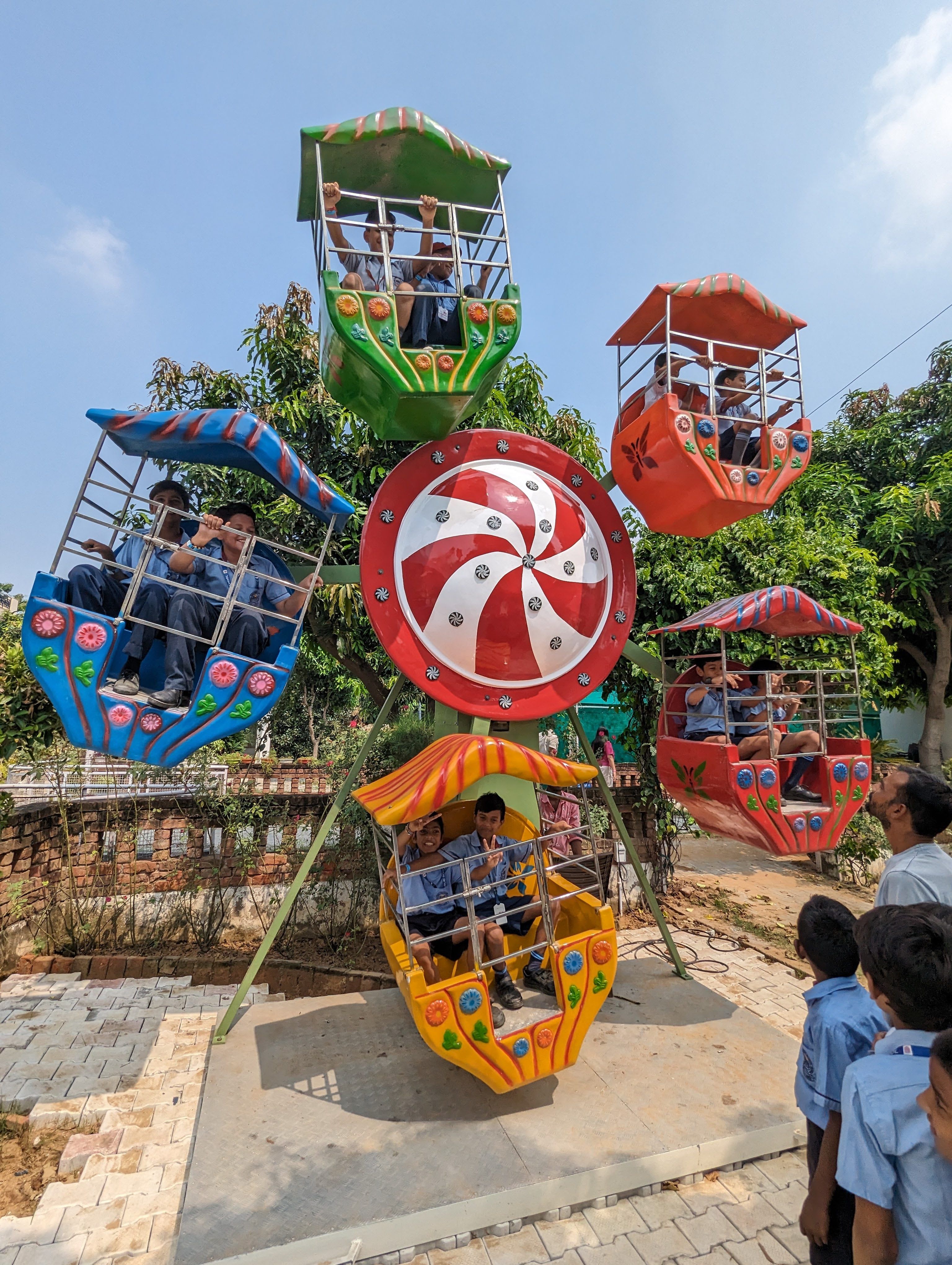 School children in uniform riding a small colourful Ferris wheel while classmates watch during a school trip at Rangmanch Farms in Gurugram near Delhi