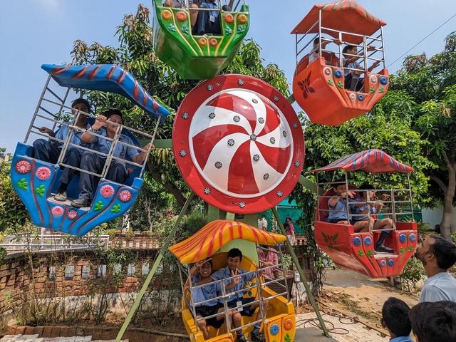 School children in uniform riding a small colourful Ferris wheel while classmates watch during a school trip at Rangmanch Farms in Gurugram near Delhi