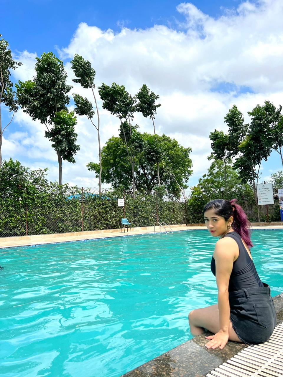 A woman seated at the edge of the outdoor swimming pool with tall trees behind, at Rangmanch Farms, a day outing and overnight stay venue in Gurugram near Delhi
