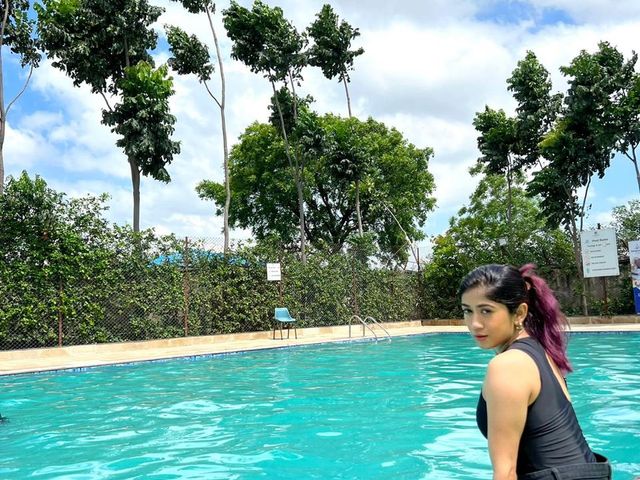 A woman seated at the edge of the outdoor swimming pool with tall trees behind, at Rangmanch Farms, a day outing and overnight stay venue in Gurugram near Delhi