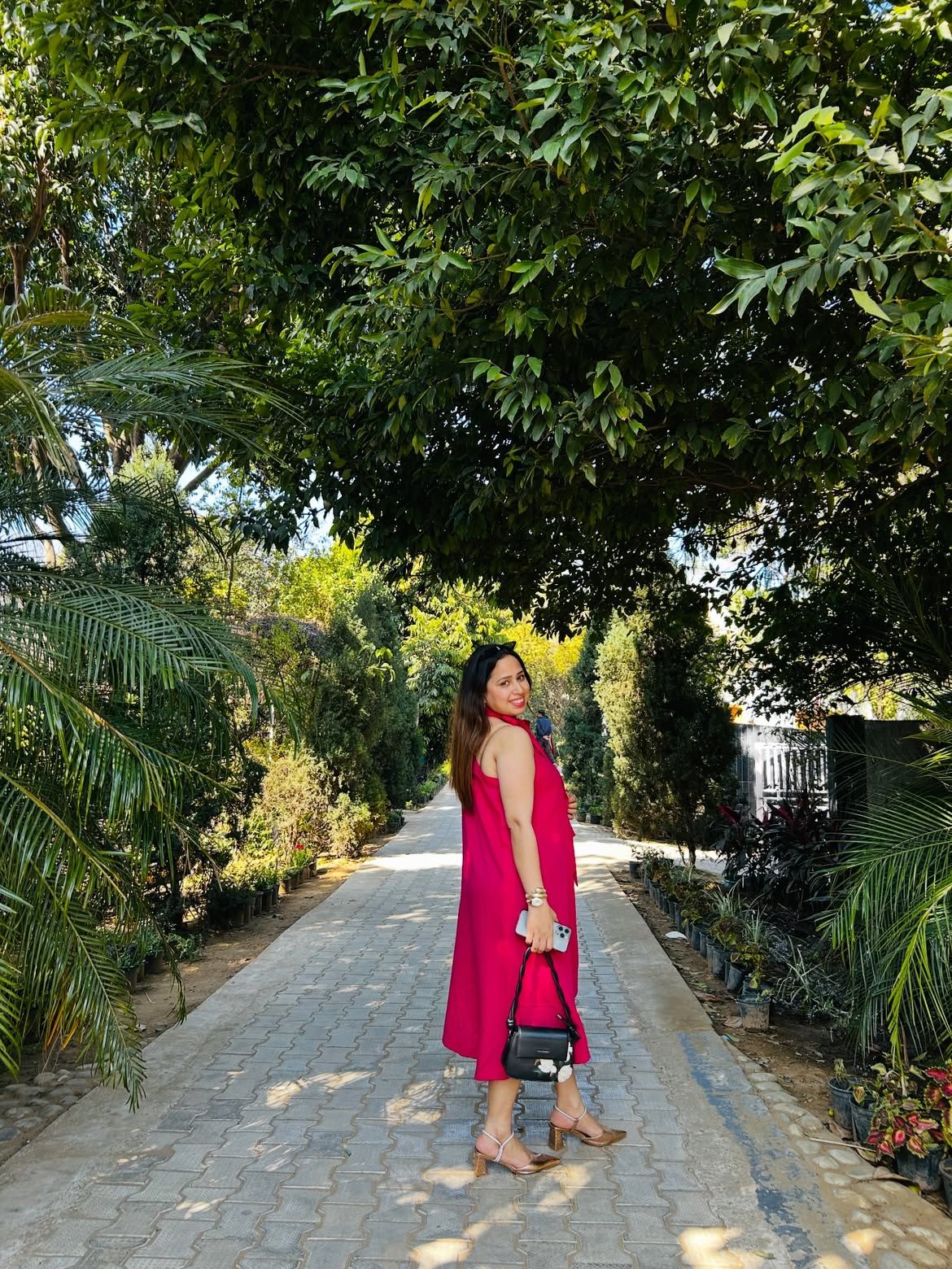 Woman in a pink dress on a paved tree-lined pathway with potted plants at Rangmanch Farms, an outdoor day outing venue in Gurugram near Delhi