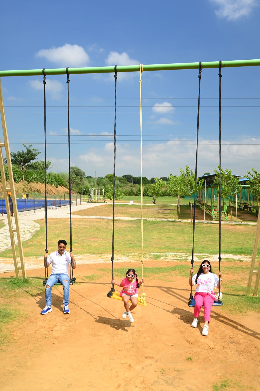 A family sitting on outdoor rope swings on the open grounds with a pool and seating area behind at Rangmanch Farms, a family day outing venue in Gurugram near Delhi