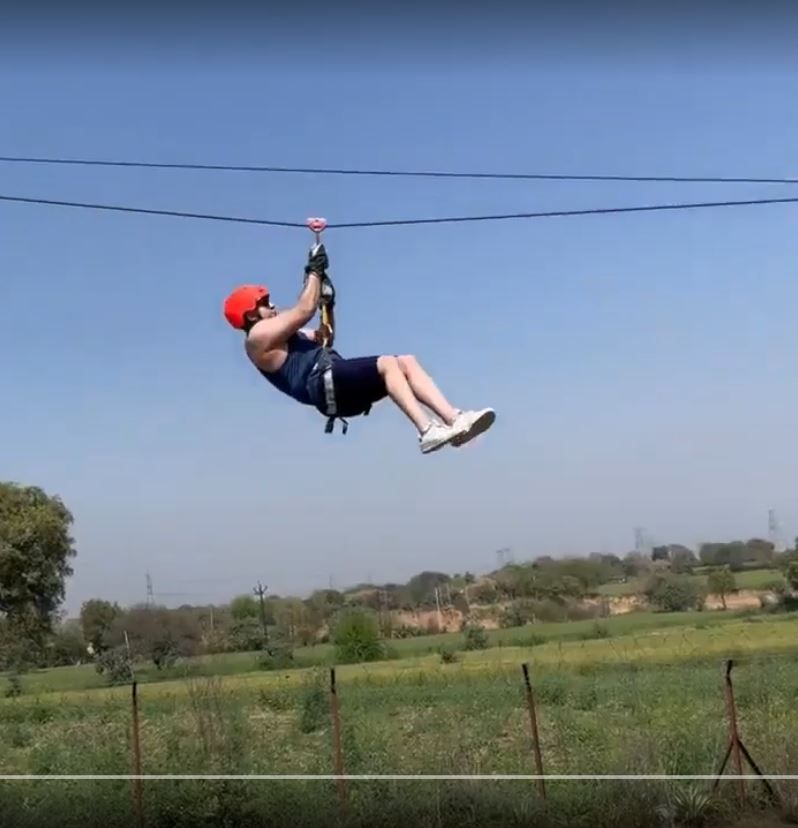 A guest in a red helmet and harness riding the zipline above green fields under a clear sky at Rangmanch Farms, an outdoor adventure and team outing venue in Gurugram near Delhi