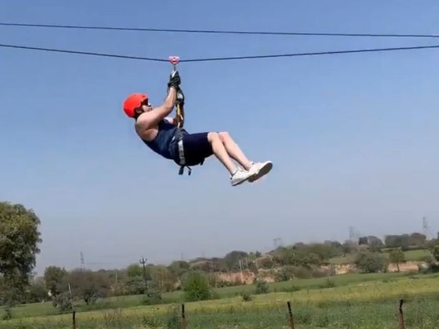 A guest in a red helmet and harness riding the zipline above green fields under a clear sky at Rangmanch Farms, an outdoor adventure and team outing venue in Gurugram near Delhi