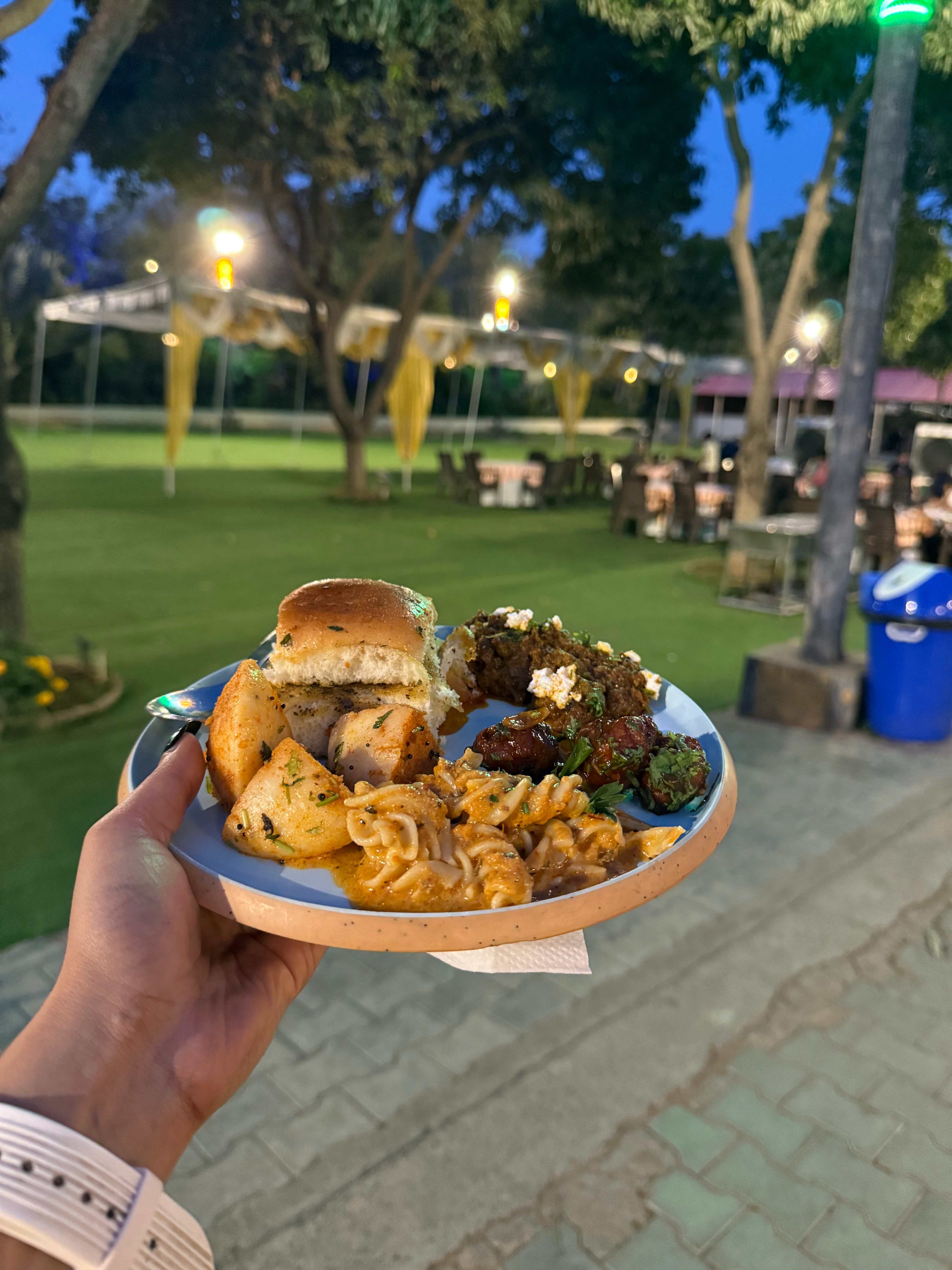 A hand holding a plate of buffet food including pasta, bread and starters at an evening outing on the lawns of Rangmanch Farms, an outdoor event venue in Gurugram near Delhi