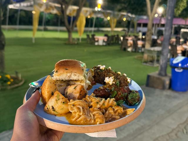 A hand holding a plate of buffet food including pasta, bread and starters at an evening outing on the lawns of Rangmanch Farms, an outdoor event venue in Gurugram near Delhi