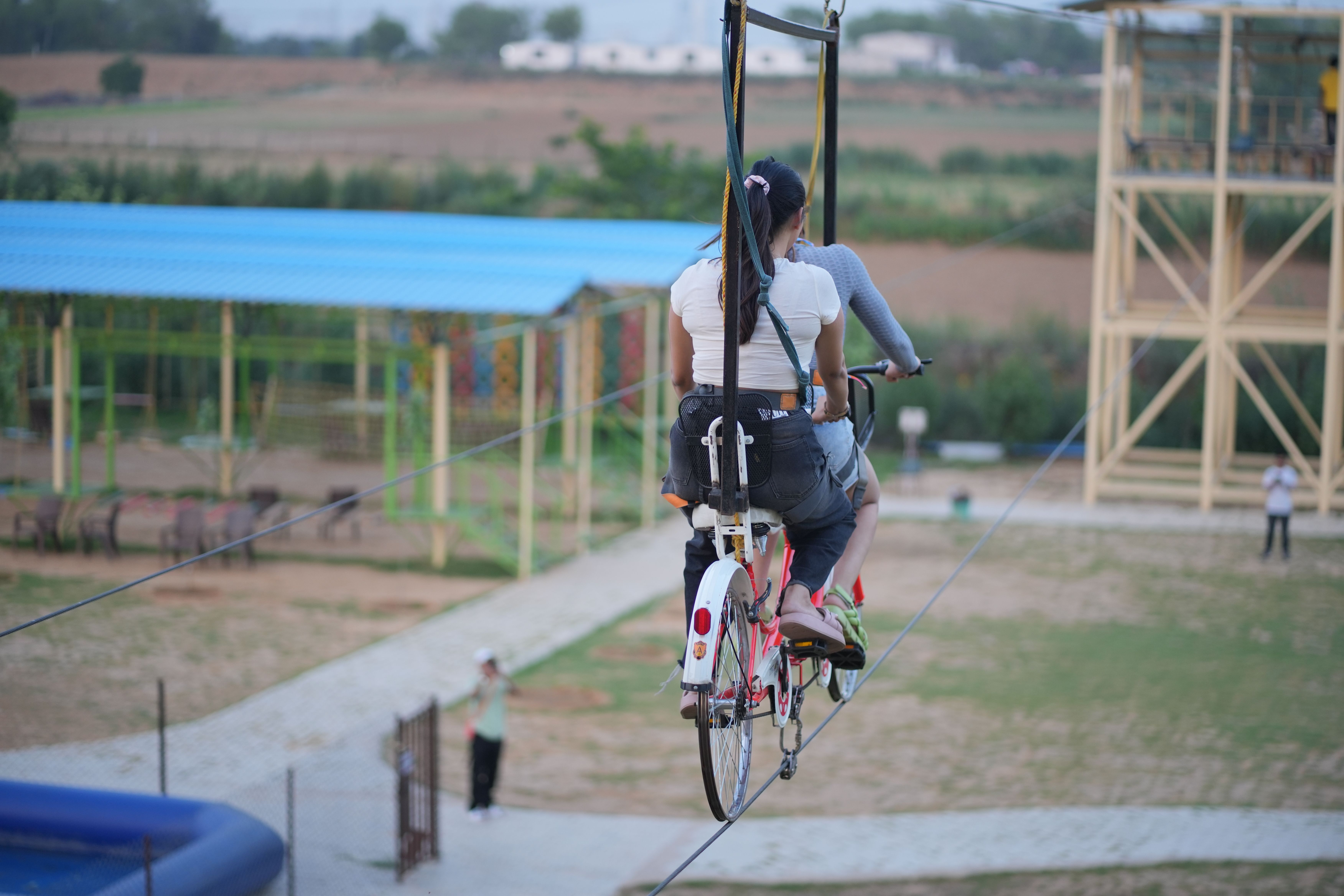 Two people riding a cycling zipline on a high rope across the outdoor adventure area at Rangmanch Farms, an outdoor day outing and team building venue in Gurugram near Delhi