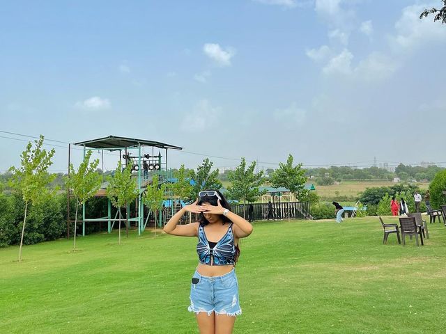 Woman standing on the open green lawn with an adventure obstacle tower behind her at Rangmanch Farms, an outdoor day outing venue in Gurugram near Delhi