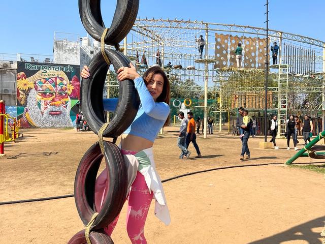 A visitor posing on a hanging tyre obstacle with the rope course and mural behind on the outdoor adventure field at Rangmanch Farms, Gurugram near Delhi