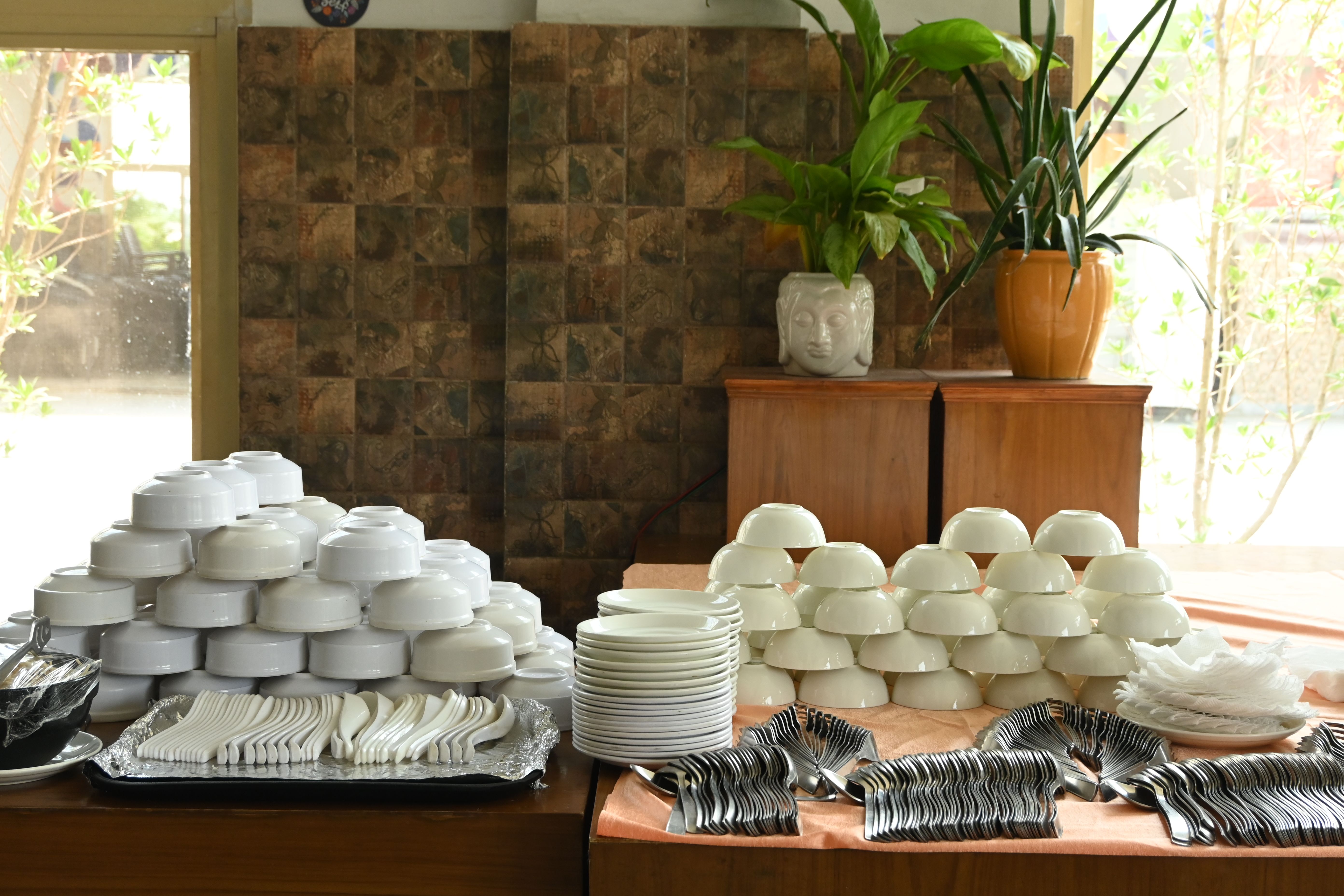 Stacked white bowls, plates, spoons and forks arranged on a buffet table with potted plants behind at Rangmanch Farms, an event and outing venue in Gurugram near Delhi