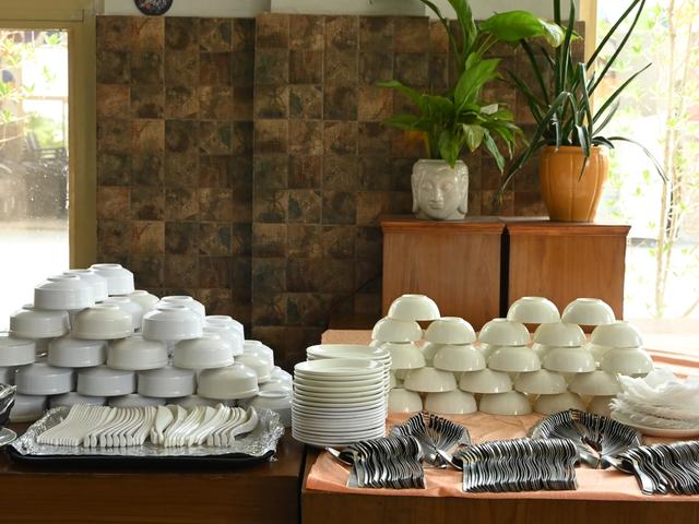 Stacked white bowls, plates, spoons and forks arranged on a buffet table with potted plants behind at Rangmanch Farms, an event and outing venue in Gurugram near Delhi