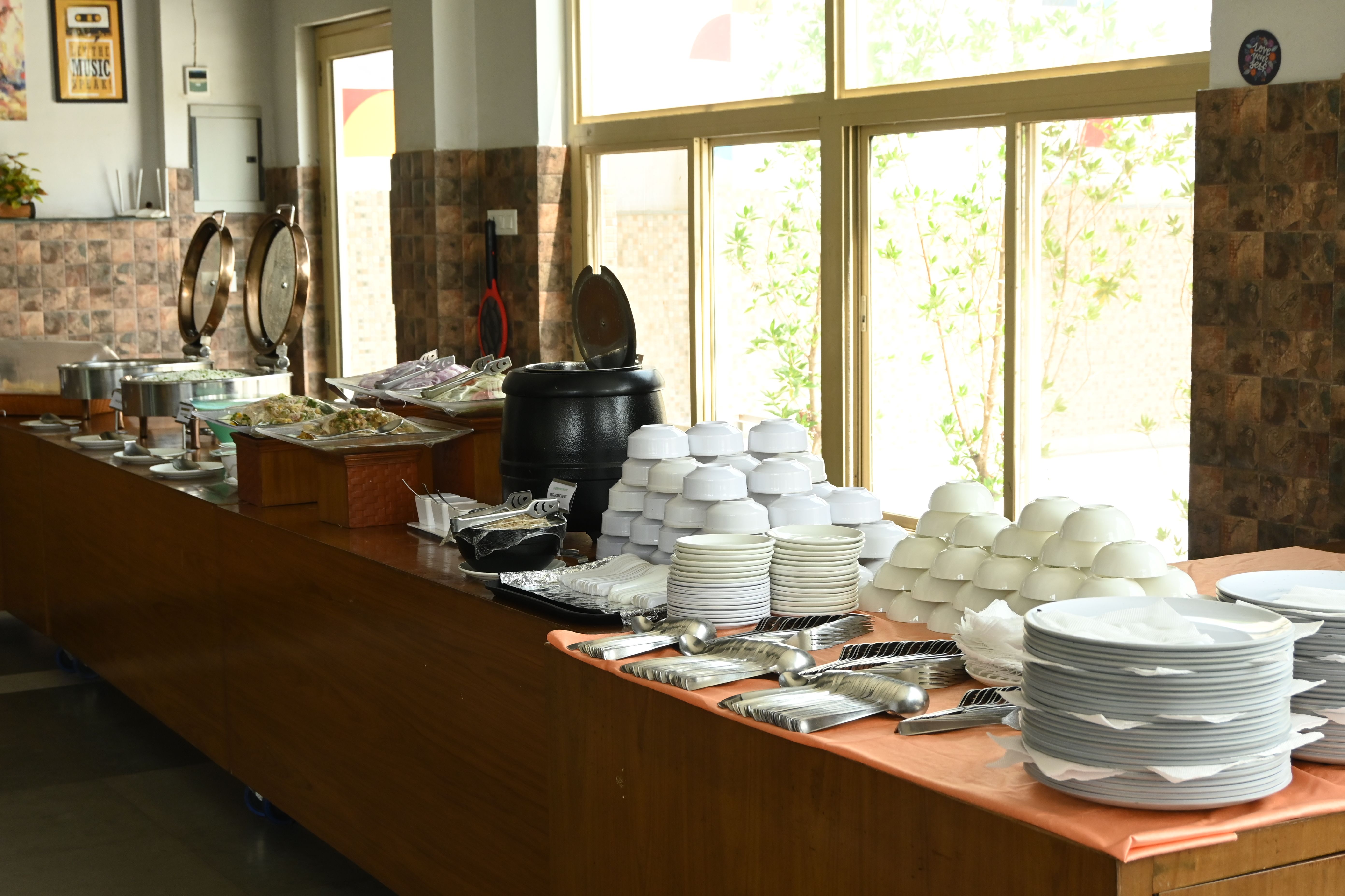 Indoor buffet counter with chafing dishes, stacked plates, bowls and cutlery laid out for an event at Rangmanch Farms in Gurugram, near Delhi
