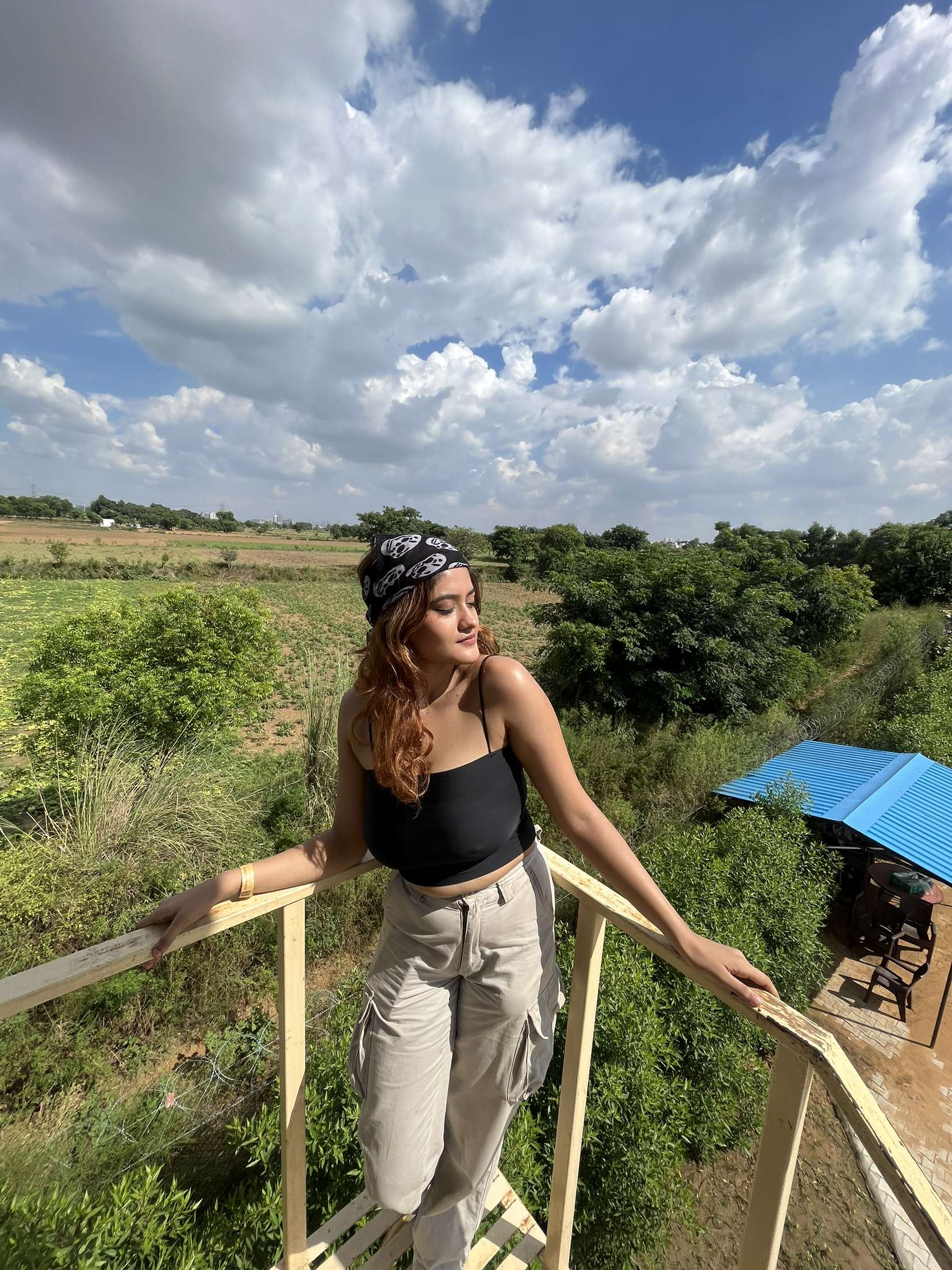 A woman posing by a wooden railing overlooking green fields and a blue-roofed shelter under a cloudy sky at Rangmanch Farms, an outdoor day outing venue in Gurugram near Delhi