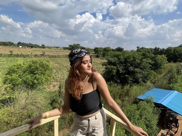 A woman posing by a wooden railing overlooking green fields and a blue-roofed shelter under a cloudy sky at Rangmanch Farms, an outdoor day outing venue in Gurugram near Delhi