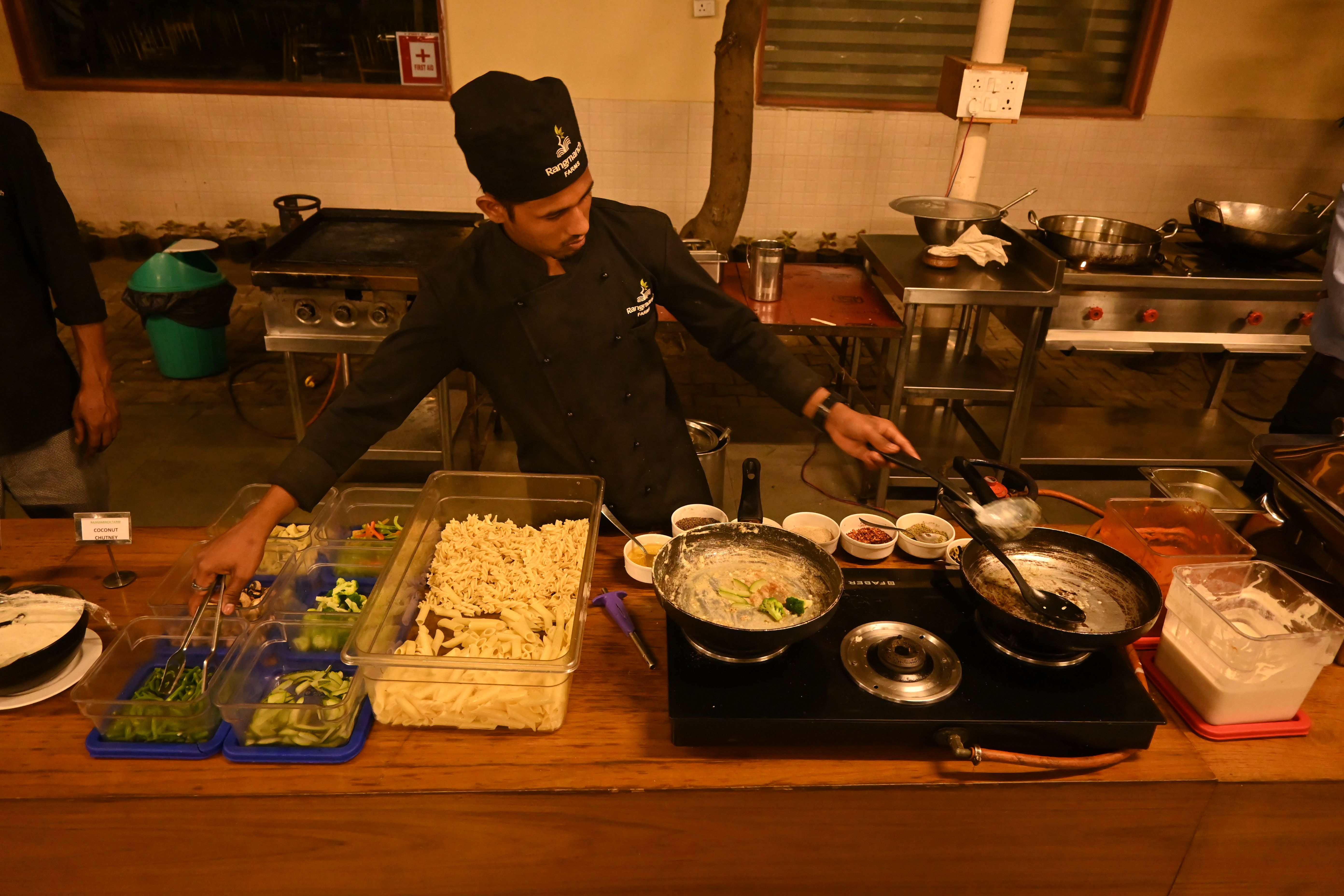 Chef preparing pasta at a live cooking counter with fresh vegetables and pans during an event at Rangmanch Farms, an outdoor venue in Gurugram near Delhi