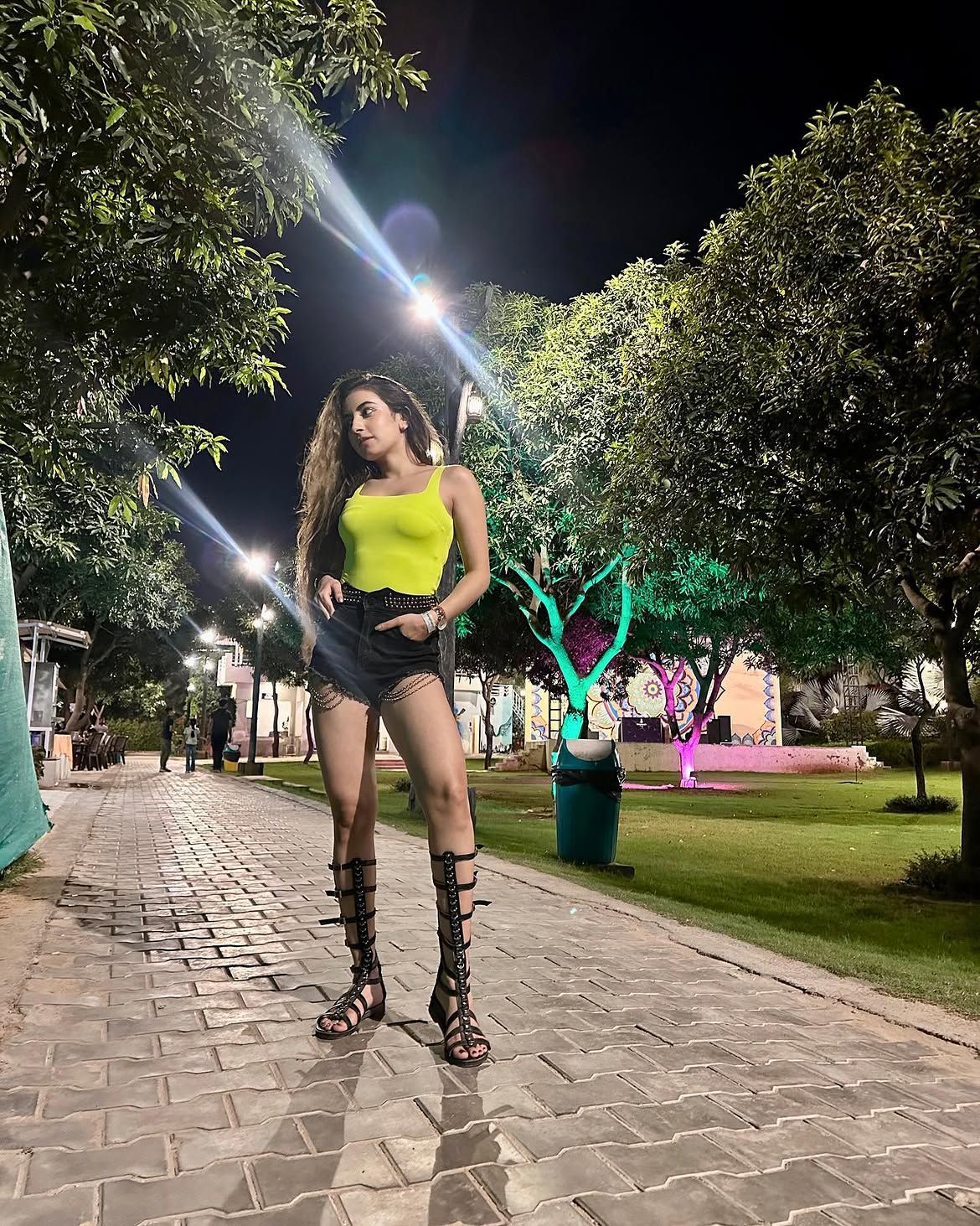 A woman posing on a tree-lined paved pathway with colourfully lit trees during an evening outing at Rangmanch Farms in Gurugram, near Delhi