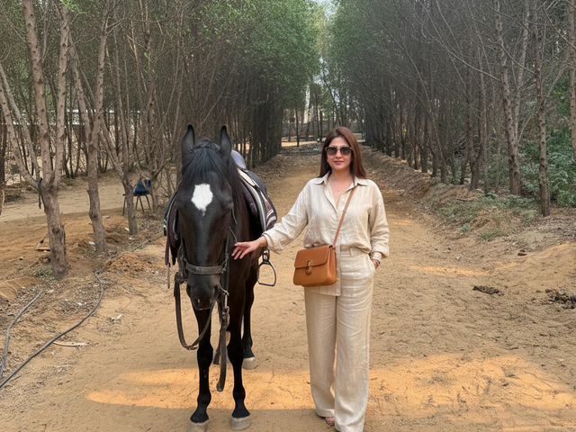 Woman standing beside a saddled black horse on a sandy tree-lined path during a day outing at Rangmanch Farms in Gurugram, near Delhi