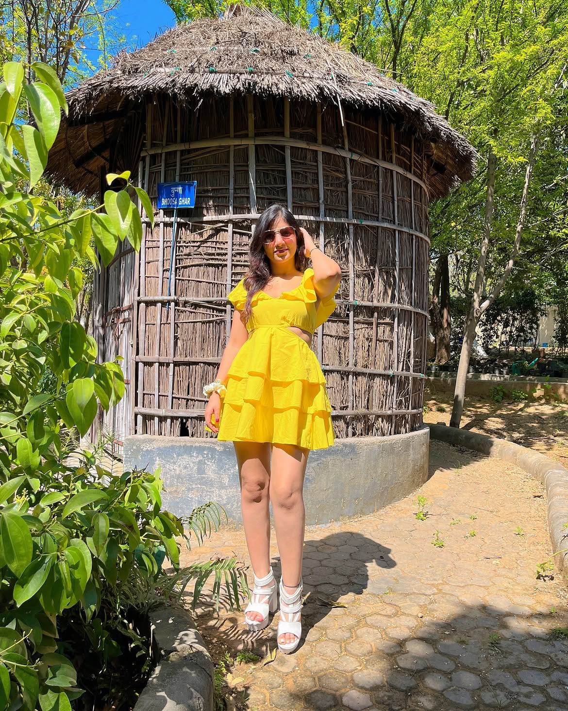 Woman in a yellow dress posing beside a round thatched mud hut surrounded by greenery at Rangmanch Farms, an outdoor day outing venue in Gurugram near Delhi