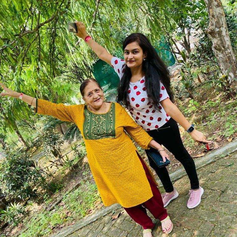 Two women posing for a selfie with arms outstretched on a tree-lined garden path during a family day outing at Rangmanch Farms in Gurugram, near Delhi