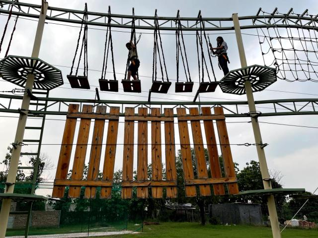 Two people harnessed on a high ropes obstacle course with hanging wooden platforms, an outdoor adventure and team building activity at Rangmanch Farms in Gurugram near Delhi