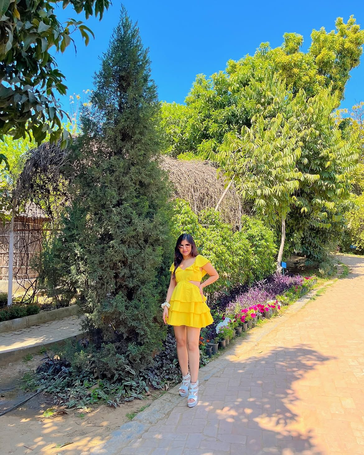 Woman in a yellow dress posing on a paved garden pathway lined with flowering plants and tall trees at Rangmanch Farms, an outdoor day outing venue in Gurugram near Delhi