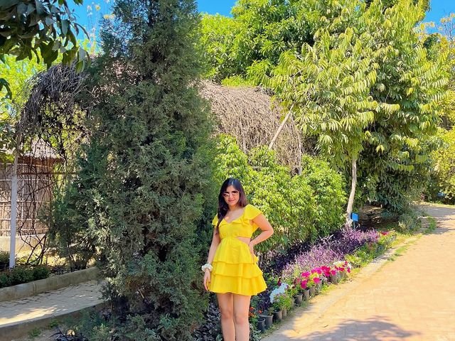 Woman in a yellow dress posing on a paved garden pathway lined with flowering plants and tall trees at Rangmanch Farms, an outdoor day outing venue in Gurugram near Delhi