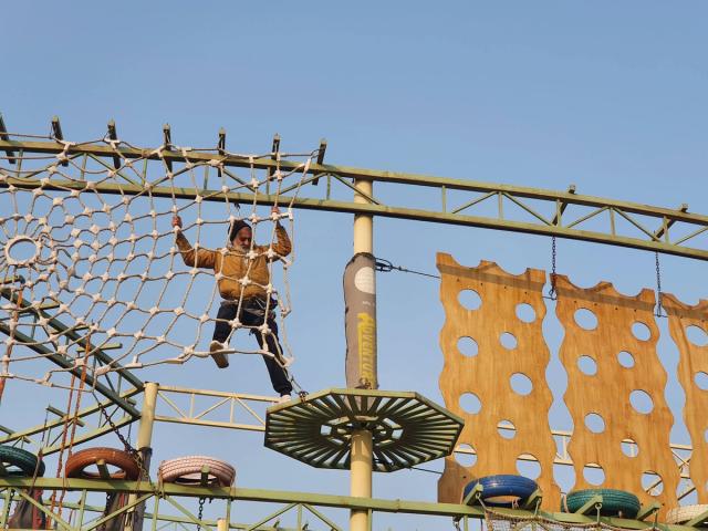 Person in a safety harness climbing a rope net on the high ropes obstacle course under a clear sky — adventure and team building activity at Rangmanch Farms in Gurugram, near Delhi