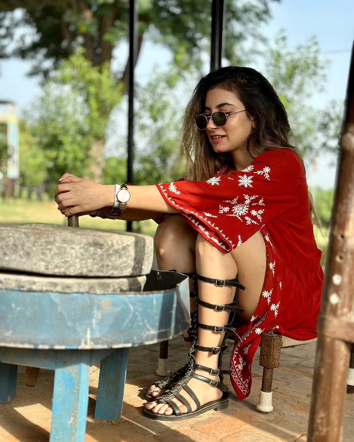 Woman in a red embroidered dress using a traditional stone hand mill during a day outing at Rangmanch Farms, an outdoor venue in Gurugram near Delhi