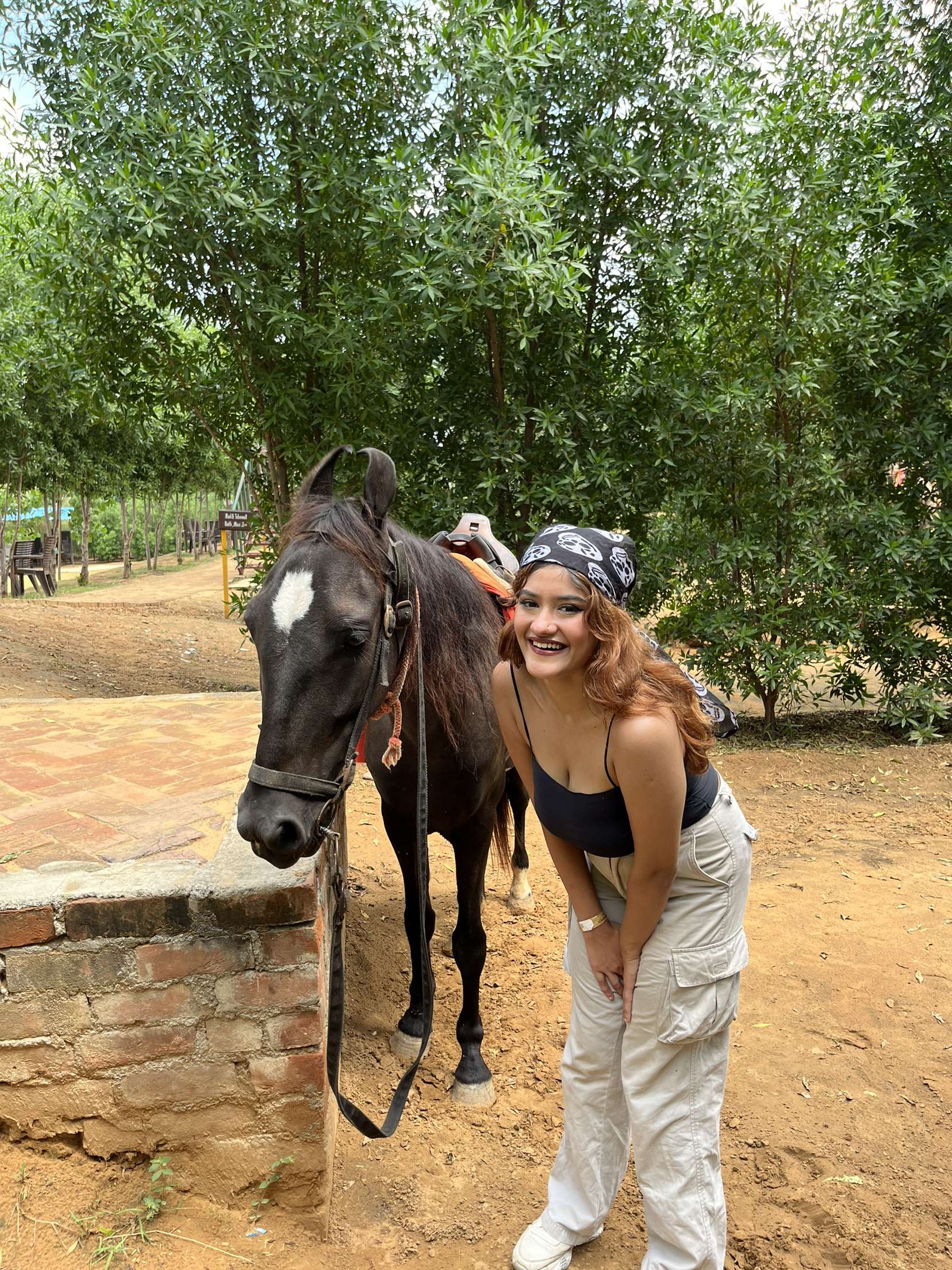 A smiling guest posing beside a saddled horse under shady trees during a day outing at Rangmanch Farms in Gurugram, near Delhi