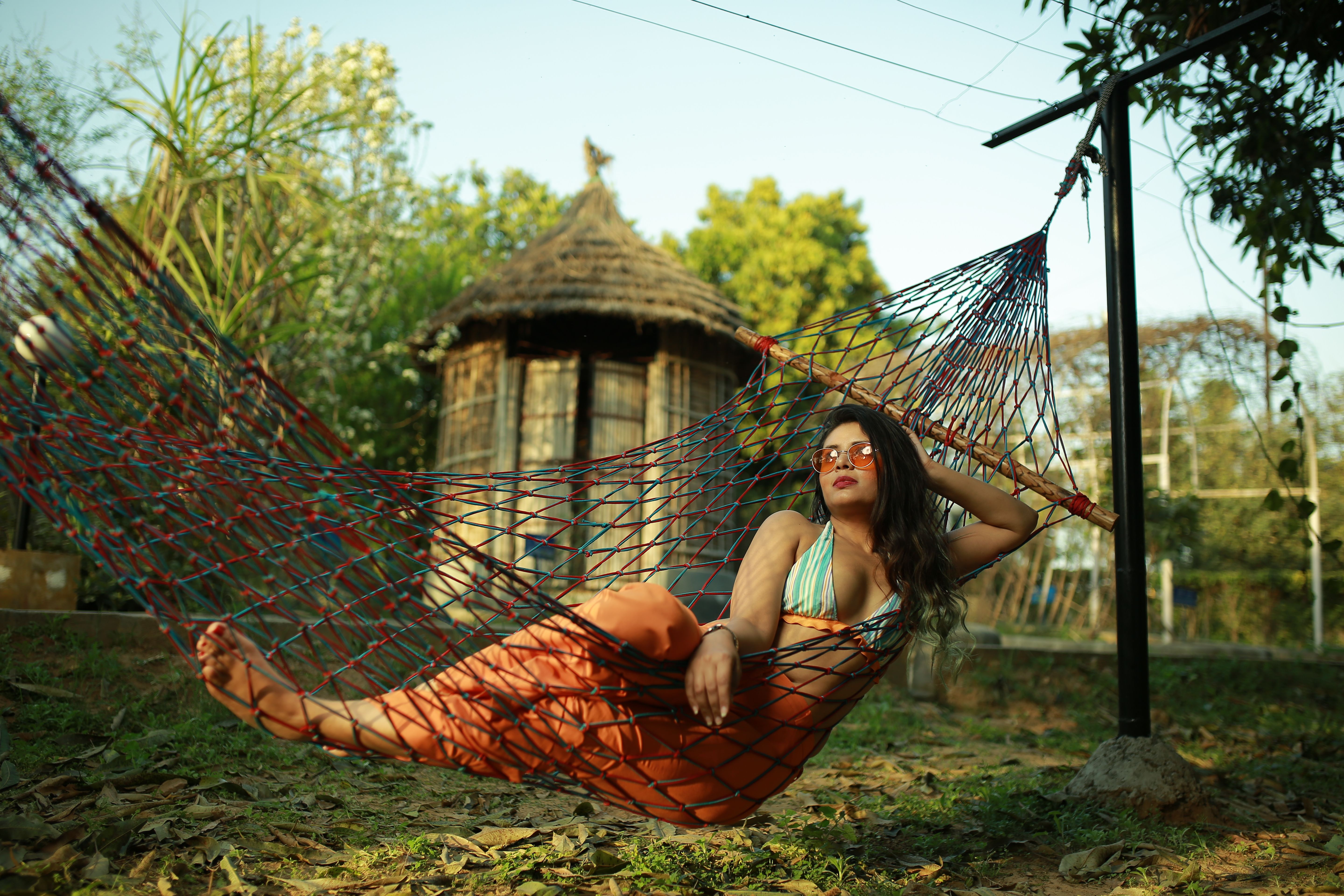 Woman in sunglasses lounging on a colourful net hammock beside a thatched hut on the grounds of Rangmanch Farms, an outdoor day outing and overnight stay venue in Gurugram near Delhi