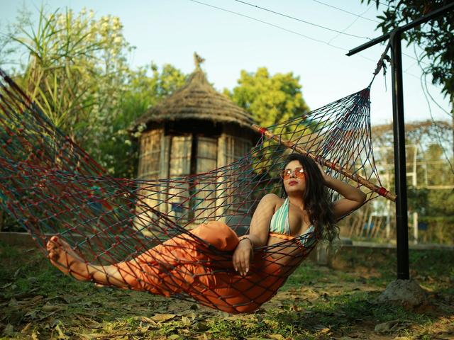 Woman in sunglasses lounging on a colourful net hammock beside a thatched hut on the grounds of Rangmanch Farms, an outdoor day outing and overnight stay venue in Gurugram near Delhi