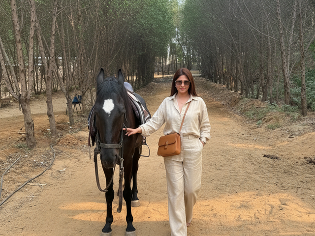 Woman standing with a saddled black horse on a tree-lined dirt trail during a day outing at Rangmanch Farms, an outdoor venue in Gurugram near Delhi