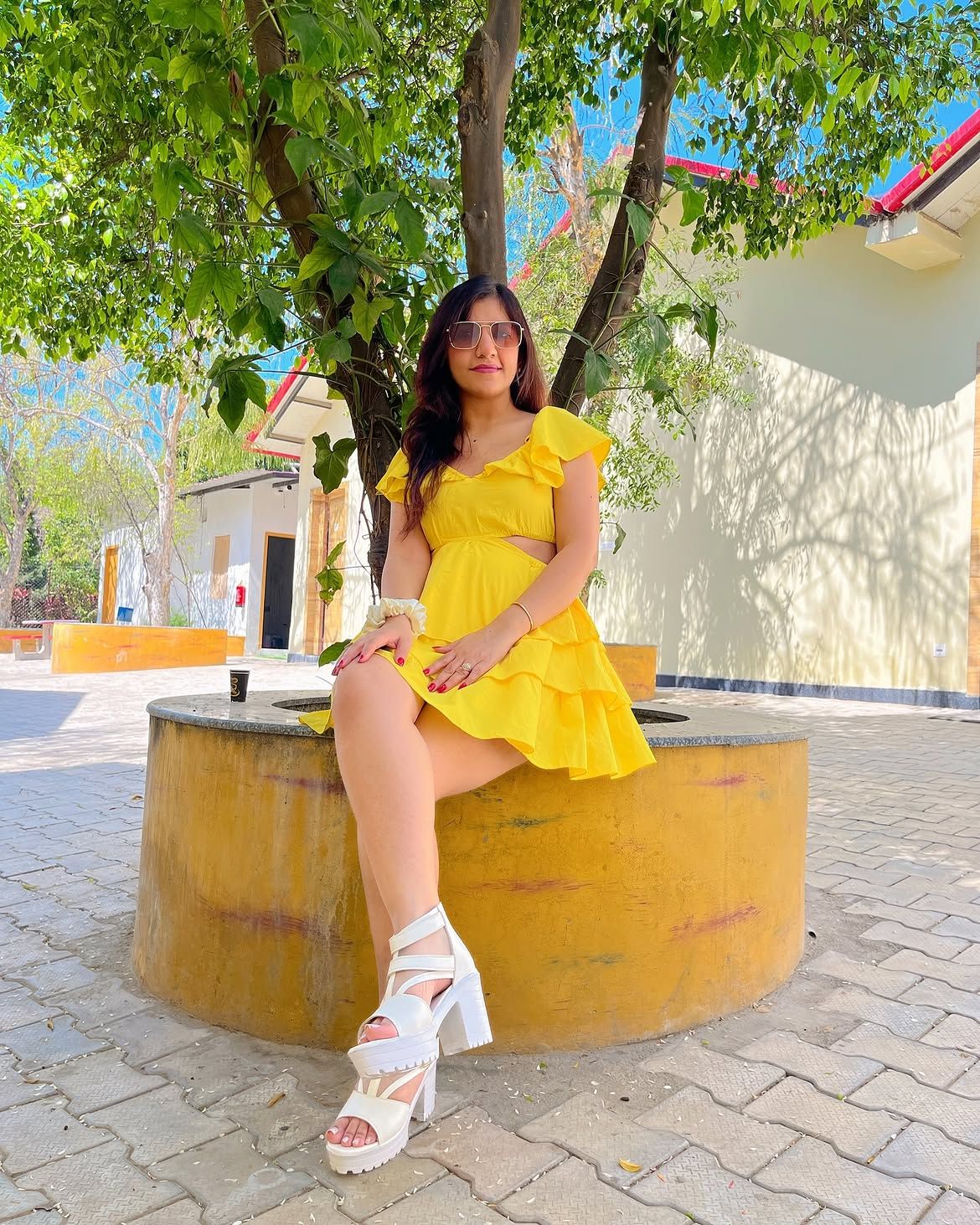 Woman in a yellow dress and sunglasses seated on a circular tree planter in a paved courtyard during a day outing at Rangmanch Farms in Gurugram, near Delhi
