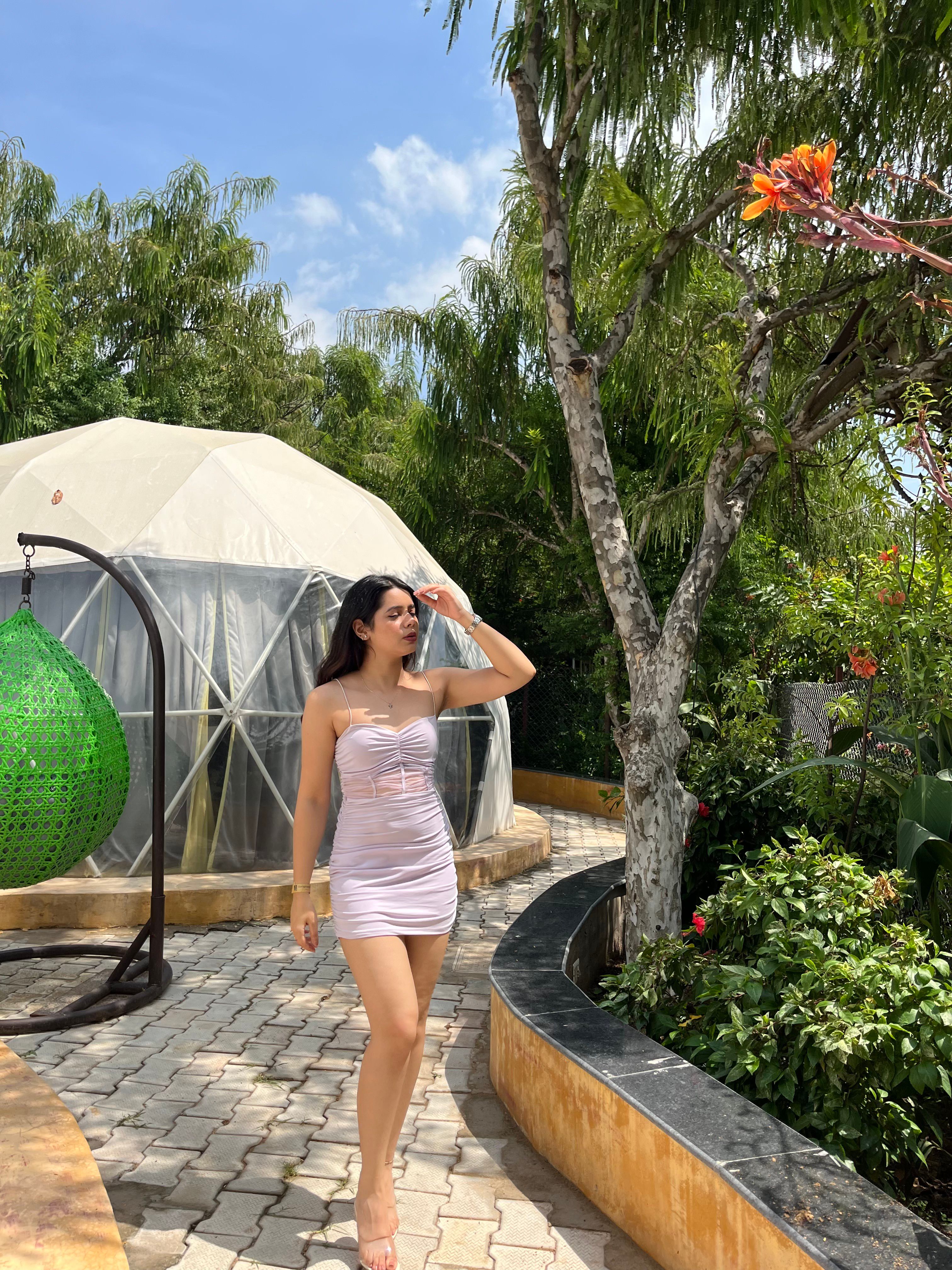 Woman in a pink dress standing beside a geodesic glamping dome and green hanging chair among trees at Rangmanch Farms, an outdoor stay venue in Gurugram near Delhi