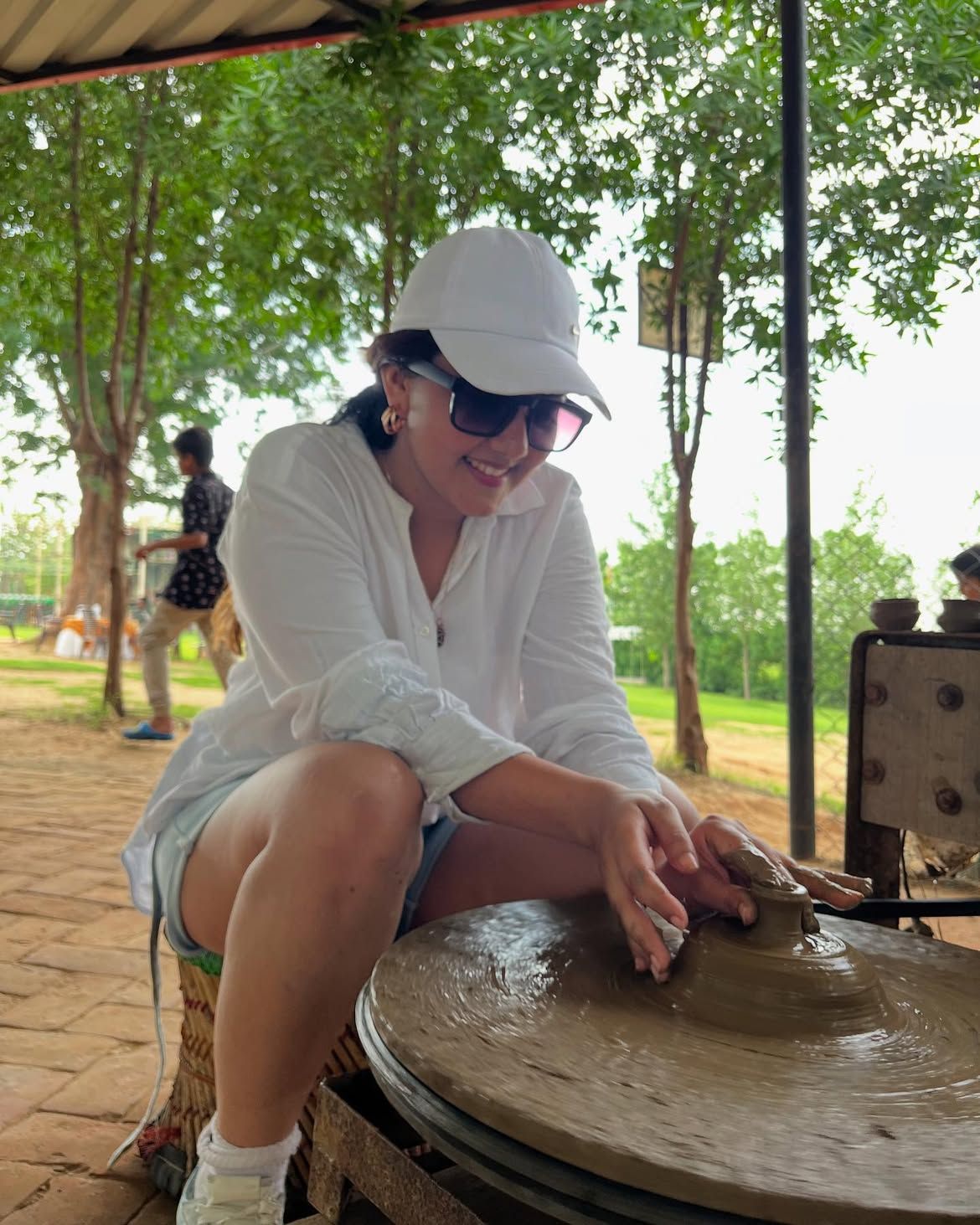 A smiling guest in a cap and sunglasses shaping wet clay at a pottery wheel during an outdoor activity at Rangmanch Farms, Gurugram near Delhi