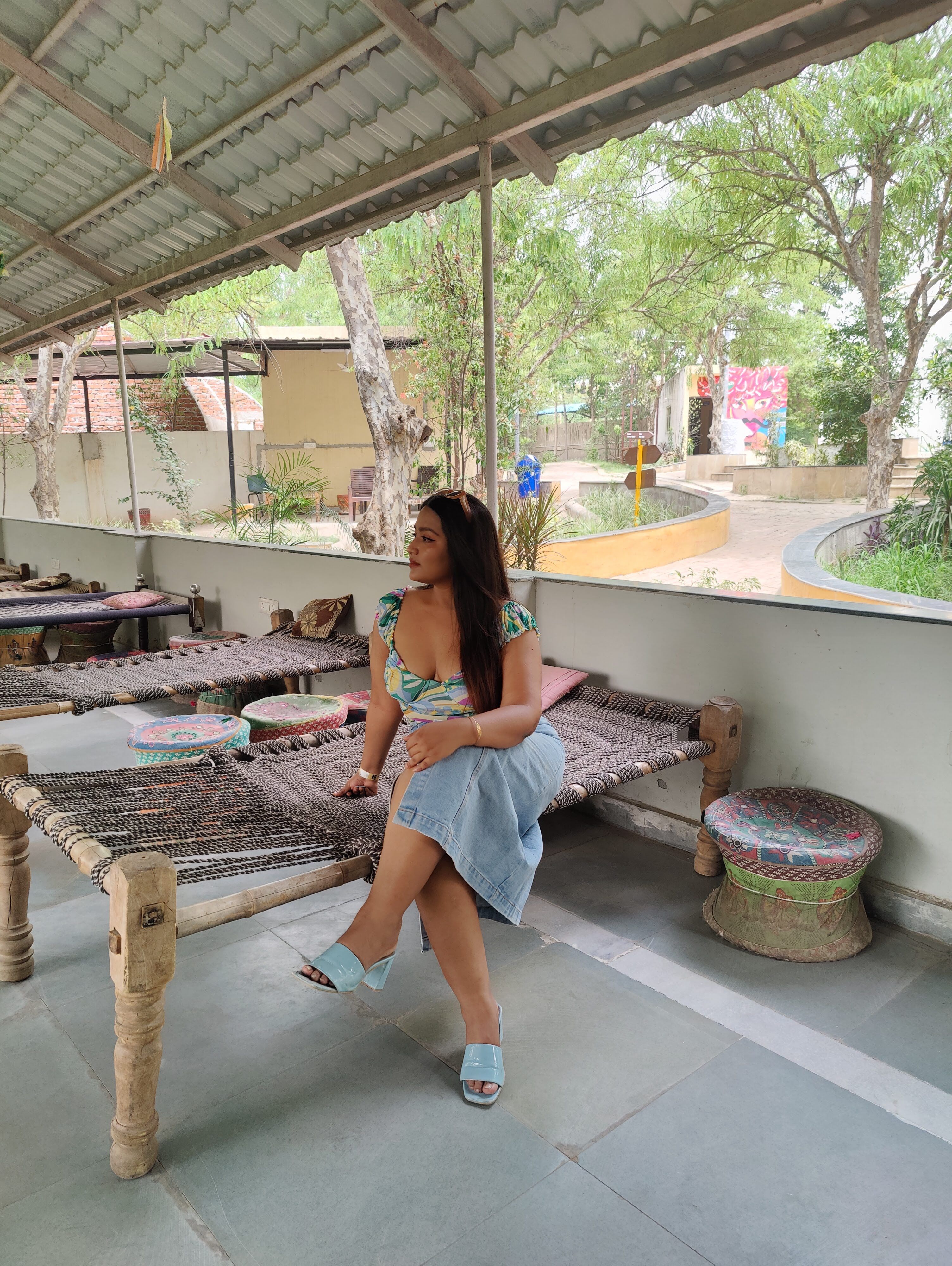 Woman seated on a traditional woven charpai in a shaded rustic seating area overlooking the greenery at Rangmanch Farms, an outdoor day outing venue in Gurugram near Delhi